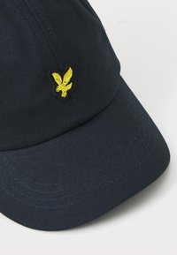 Căciulă de culoare albastru marin, confecționată dintr-un material texturat, cu o margine curbată. Prezintă un logo brodat în galben al unei vulturi pe partea din stânga față.
