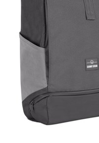 Rucsac gri cu un material texturat, accente netede, buzunar lateral și un etichetă mică personalizată cu "JOHNNY URBAN."