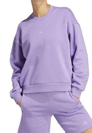 Person iført en matchende lavendelfarvet Adidas sweatshirt og shorts med hænderne i lommerne, intet hoved synligt, på en hvid baggrund.