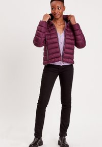 Doudoune maroon avec fermeture éclair, poches inclinées et capuche. Portée par-dessus un pull lilas clair et associée à un jean noir et des bottines.