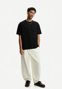 Črna teksturirana kratkotrajna srajca v kombinaciji z belimi širokimi hlačami in črnimi čipkastimi čevlji, ki jih dopolnjuje minimalistična srebrna verižica.