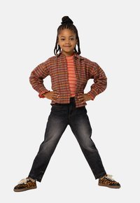 Plaid jacket in orange and red over a red striped shirt, with dark jeans and leopard print sneakers. Child stands confidently with hands on hips.