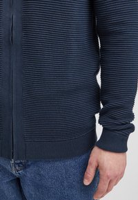 Granatowy sweter na zamek błyskawiczny z prążkowaną fakturą, długimi rękawami i dopasowanym dołem. W połączeniu z niebieskimi dżinsami, pokazującą dłoń blisko mankietu swetra.