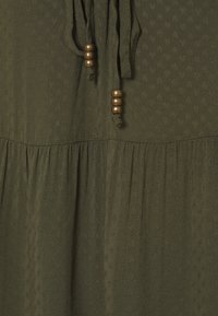 Túnica verde oliva con tejido texturizado, que presenta un escote anudado y acentos de cuentas de madera. Dobladillo acampanado con detalle sutil de patrón en todo su diseño.