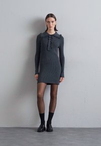 Knit, long-sleeve, dark grey dress with a ruffled collar, button detail, fitted design, and solid color. Model wears black tights and shoes.