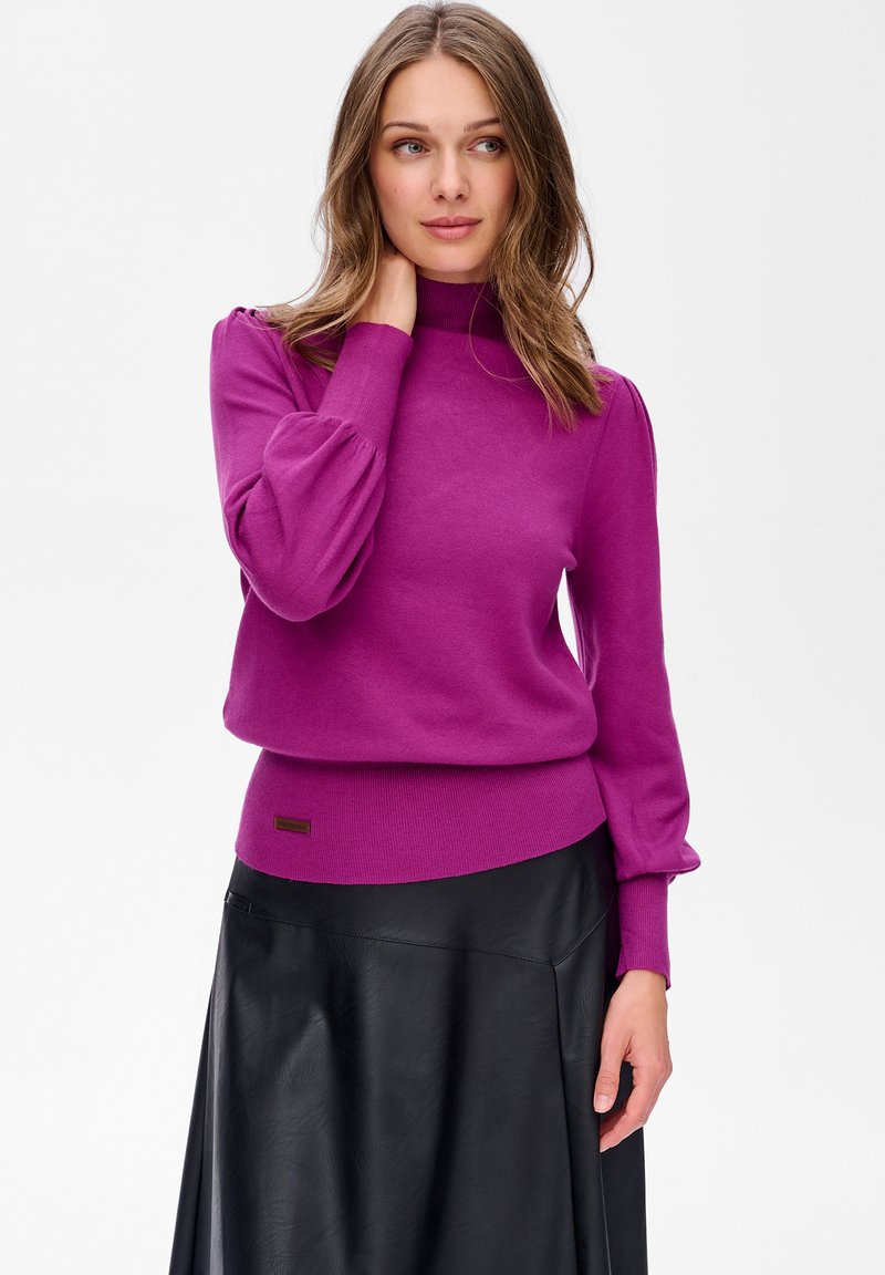 Kobieta o jasnobrązowych włosach, ubrana w fioletowy sweter z długimi rękawami i czarną skórzaną spódnicę, pozująca z jedną ręką dotykającą szyi, patrząca w prawo.