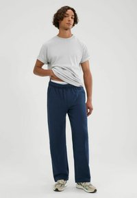 Graues Baumwoll-T-Shirt mit kurzen Ärmeln kombiniert mit einer schwazen Hose mit elastischem Bund. Das Model trägt beigen und grünen Sneakers mit Muster.