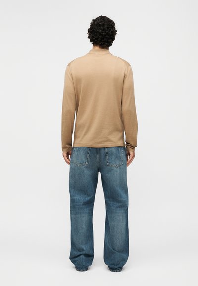 Άνδρας με σγουρά μαλλιά στέκεται με την πλάτη, φοράει ένα μπεζ μακρυμάνικο πουκάμισο και φαρδιά μπλε τζιν σε ένα απλό φωτεινό φόντο.