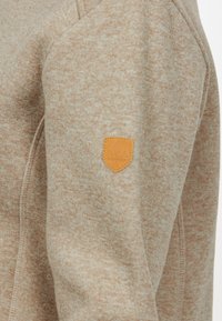 Beige stickad sweatshirt med en texturerad finish. Har en liten, brun läderlogotyp på ärmen. Klassisk design med rund hals.