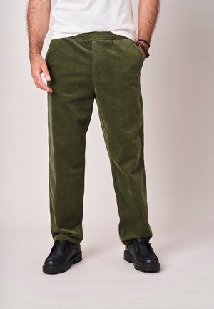 Man draagt olijfgroene corduroy broek, zwarte schoenen en wit t-shirt, staat met één hand in de zak en grijze achtergrond.