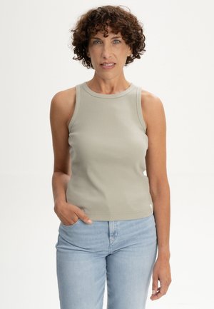 Vrouw met krullend bruin haar, gekleed in een mouwloos beige topje en lichtblauwe spijkerbroek, staand met één hand in haar zak.