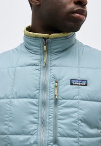 Άνδρας με ανοιχτό μπλε μπουφάν Patagonia με κουμπιά σε κίτρινο φερμουάρ, στραμμένος προς τα δεξιά, φαίνεται από τον λαιμό έως το στήθος.