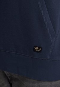 Marineblauwe sweatshirt met een gladde textuur, voorzien van een zwart label met gouden tekst nabij de zoom en contrasterende stiksels aan de zijkant.