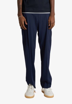 Persona che indossa pantaloni cargo blu navy larghi, sneaker bianche con suola in gomma e una maglietta nera con orlo bianco, in piedi davanti a uno sfondo semplice.