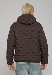 Brun boblejakke med et teksturert, quiltet design og hette. Har en figurnær silhuett og ribbestrikkede mansjetter, kombinert med lyseblå jeans.