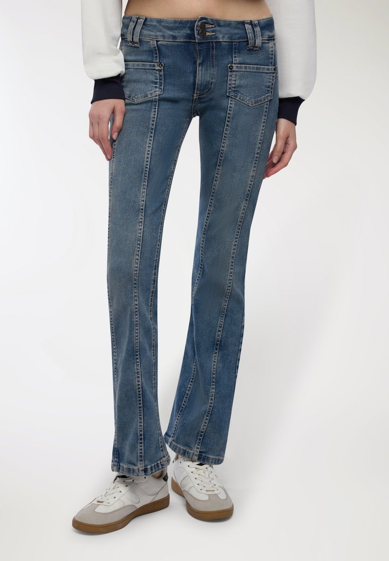 Donna che indossa jeans in denim blu con tasche frontali e sneakers bianche, in piedi contro uno sfondo chiaro e semplice.