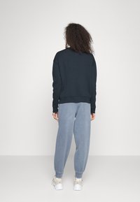 Donna con capelli ricci che indossa una felpa scura, pantaloni jogger azzurri chiari e sneakers bianche, in piedi di fronte a uno sfondo grigio chiaro uniforme.