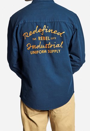 Homme portant une chemise bleu marine avec un texte orange dans le dos indiquant "Redefined Rebel 2013 Industrial Uniform Supply", associé à un pantalon beige.
