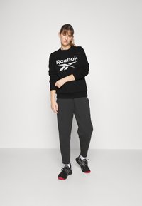 Felpa nera con logo Reebok bianco, pantaloni della tuta grigi e scarpe sportive nere con dettagli rossi. Tessuto liscio, vestibilità rilassata.