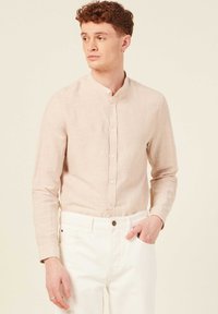 Camicia di lino color beige chiaro con colletto alla mandarino, chiusura frontale con bottoni, maniche lunghe e abbinata a pantaloni bianchi. Design semplice e pulito.