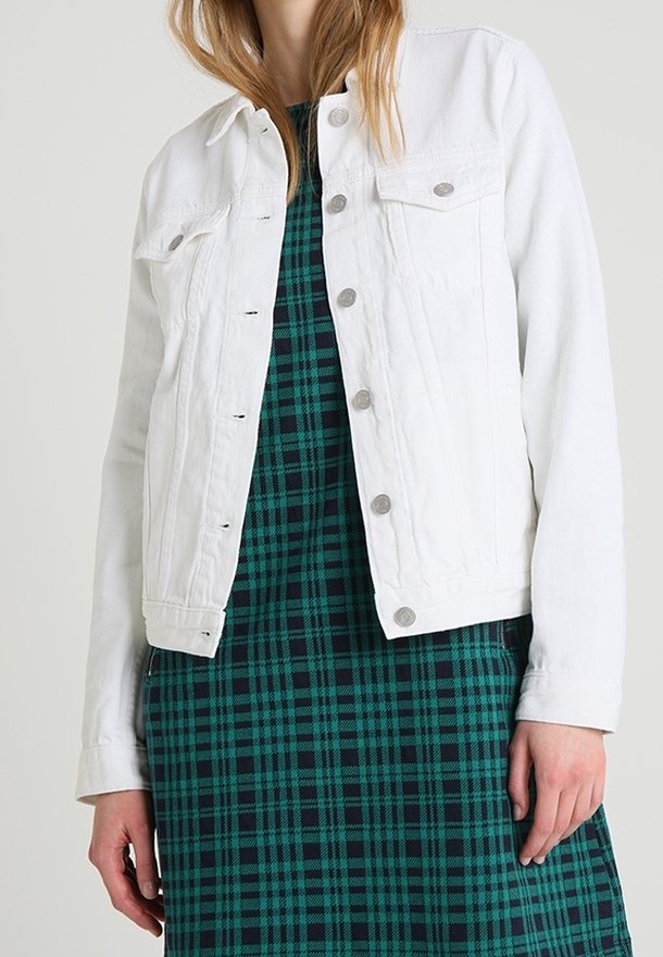 Femme portant une veste en denim blanche sur une robe à carreaux verte et marine, se tenant devant un fond uni clair.