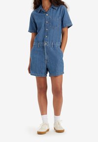 Denimový overal s krátkým rukávem, zapínáním na knoflíky a náprsní kapsou. Světle modrá barva, volný střih a ohrnuté šortky. Pár bílých tenisek.
