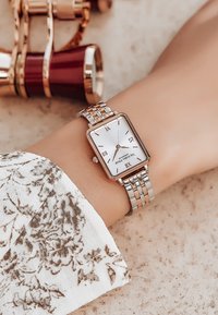 Reloj rectangular plateado con una cara blanca, números romanos negros y una pulsera metálica de eslabones en una muñeca, junto a una tela con patrones.