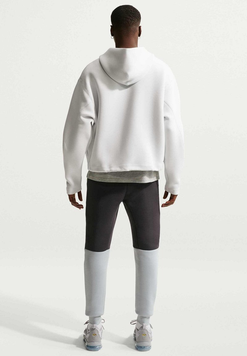 Mand med ryggen til iført en hvid hættetrøje, sorte og grå farveblok-bukser og lysegrå sneakers med gennemsigtige såler.