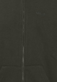 Felpa con zip verde scuro realizzata in tessuto morbido, con zip frontale, due tasche laterali e un logo discreto sul petto.