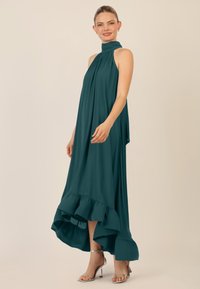 Teal halterneck maxikjole med flydende stof, høj halsudskæring og bølget kant. Har et high-low snit og en glat tekstur. Sølvhæle bæres.