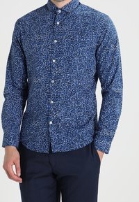Camicia blu navy con bottoni e un motivo floreale bianco. Presenta un colletto, maniche lunghe e un taglio aderente, realizzata in tessuto leggero.