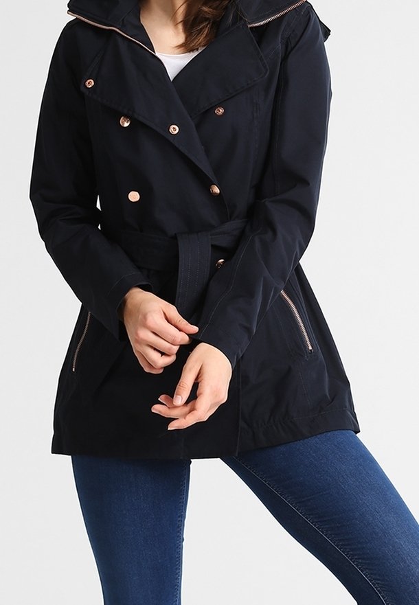 Femme portant une veste noire ceinturée avec des boutons et des fermetures éclair en or rose, associée à un jean bleu sur un fond uni.