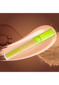 Tube transparent de correcteur Wonder Snatch avec bouchon vert citron sur fond de maquillage liquide beige avec éclaboussures de poudre et de liquide.