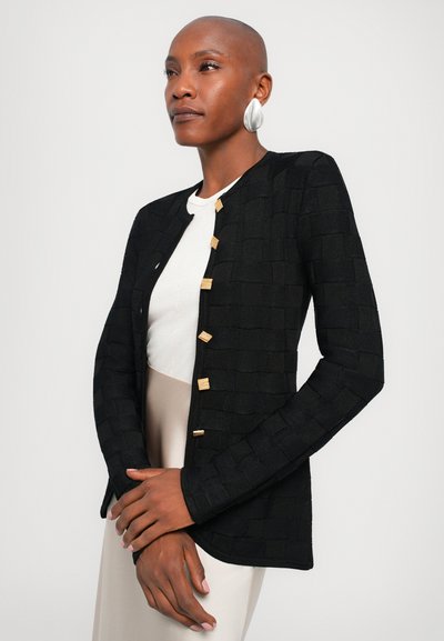 Black knitted cardigan with a textured square pattern, featuring gold buttons and a rounded neckline. Paired with a light-colored top.