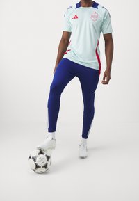 Ljusblå adidas-tröja med röda och marinblå detaljer, kombinerad med åtsittande marinblå byxor. Vita skor och en svart-vit fotboll.