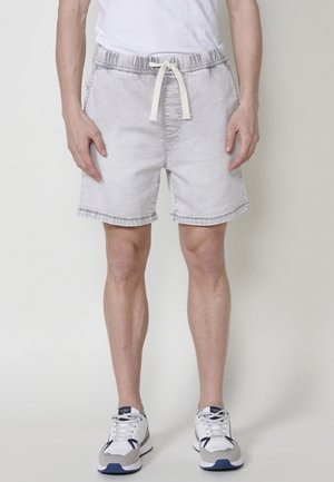 Shorts en denim gris clair avec une taille élastique et un cordon de serrage, dotés de poches latérales et d'un ourlet brut. Portés avec des baskets grises et bleu marine.
