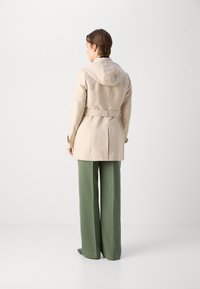 Beige trenchcoat met kap, ceintuur in de taille en rugventilatie. Draag het over groene wijde broekspijpen, met een zachte textuur en een gestructureerd ontwerp.