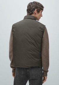 Ciemnozielona pikowana kamizelka z wysokim kołnierzem, widziana od tyłu, zestawiona z brązowym swetrem i szarymi dżinsami. Gładka faktura, minimalne detale.