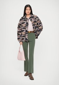 Γυναίκα ντυμένη με καμουφλάζ μπουφάν, ροζ μπλούζα, πράσινο παντελόνι, καφέ μπότες, κρατώντας μια ανοιχτό ροζ τσάντα, να στέκεται μπροστά από λευκό φόντο.