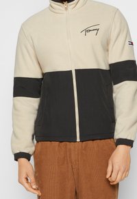 Beiget ja mustat fleecetakit, joissa on vetoketju edessä, korkea kaulus ja rintapieleen brodeerattu logo. Teksturoidusta kankaasta valmistettu, joustavat hihansuut.