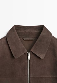 Colletto di giacca in pelle scamosciata marrone scuro con una cerniera metallica argentata parzialmente chiusa su sfondo bianco.