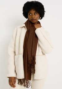 Chaqueta de forro polar crema con dos bolsillos frontales, acompañada de una bufanda marrón con detalles moteados y bordes con flecos.