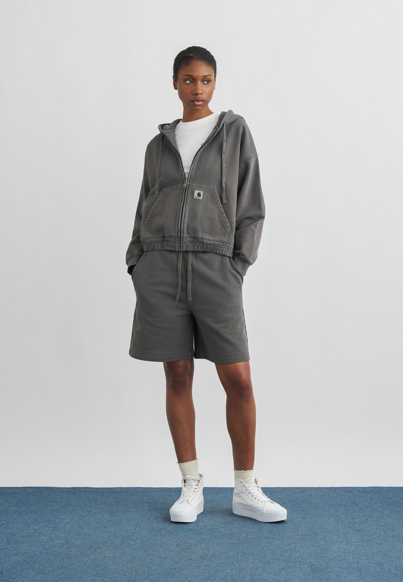 Donna con felpa grigia con zip e pantaloncini abbinati, in piedi con le mani nelle tasche su un tappeto blu contro una parete bianca.