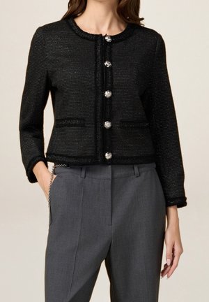 Femme portant une veste texturée noire boutonnée avec des boutons argentés et un pantalon gris taille haute à coupe ajustée, main dans la poche.