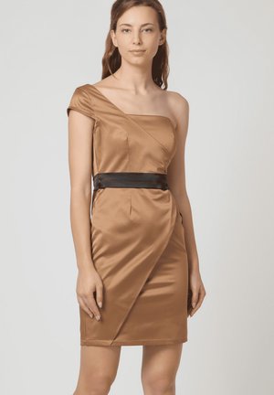 Mujer vestida con un vestido de satén bronce de un solo hombro y una cinturilla negra, de pie contra un fondo claro y liso.