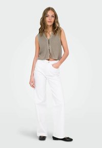 Afskåret, beige, strikket vest med bånd foran, parret med højtaljede, hvide, vide jeans og sorte balletflader, stående mod en lys baggrund.