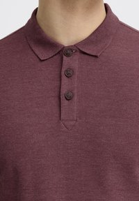 Mørkerød polo-skjorte med tekstureret overflade, der har en klassisk krave, fire brune knapper og en subtil v-formet sømdetalje ved udskæringen.
