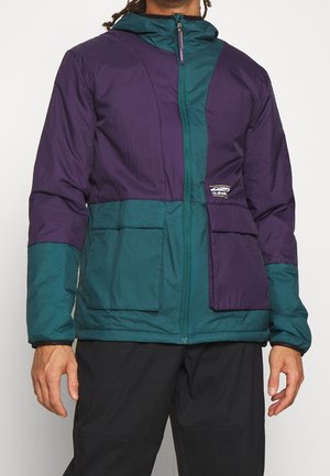 Mand iført en lynlåstrøje med farveblokke i lilla og blågrøn, med store forlommer og hætte.