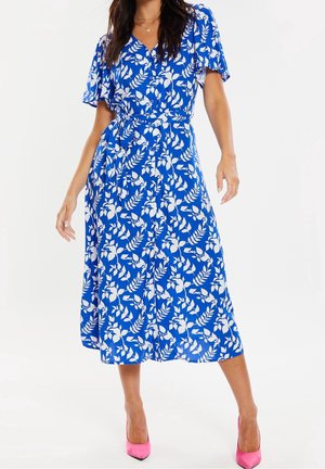 Vrouw draagt een blauwe midi wikkeljurk met witte bladpatronen en zwierige mouwen, gecombineerd met felroze hoge hakken.