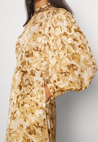 Abito con maniche a sbuffo in tessuto trasparente con un motivo floreale in tonalità oro e beige. Presenta un colletto alto e tasche laterali.